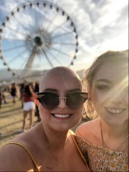 Mia, who has no hair in this photo, is at a music festival with a friend. A Ferris Wheel spins in the background. Mia wears sunglasses and smiles at the camera.