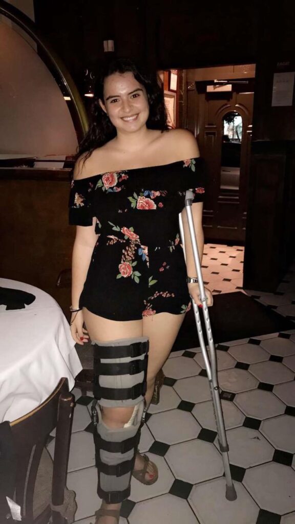 Mia stands in a black floral romper at the start of her osteosarcoma journey. She has a crutch under her left arm and a cast on her right leg.