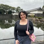 Mia Sandino has osteosarcoma. In this photo, she stands in front of some water. She is wearing a black shirt and jeans, holding a pink purse, and has curly brown hair.