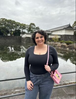 Mia Sandino has osteosarcoma. In this photo, she stands in front of some water. She is wearing a black shirt and jeans, holding a pink purse, and has curly brown hair.